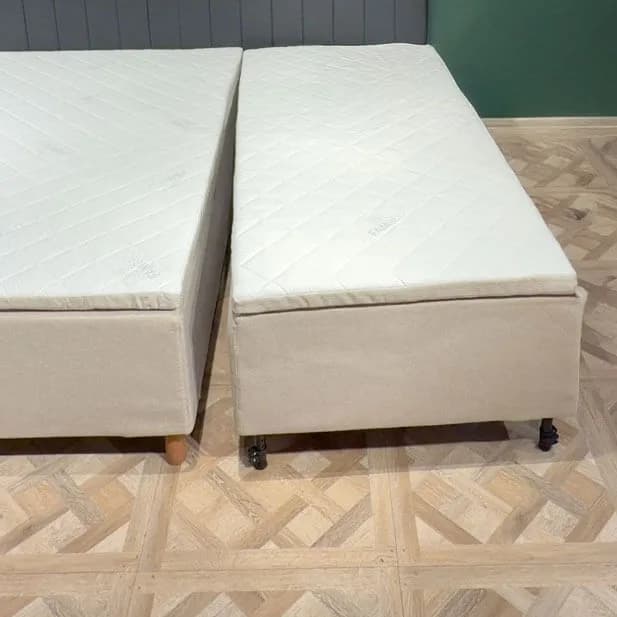To ferdige sengemoduler står ved siden av hverandre på et lyst tregulv med en smal glipe mellom. Hver modul har sitt eget beige trekk, sin egen quiltede madrass og sine egne ben.