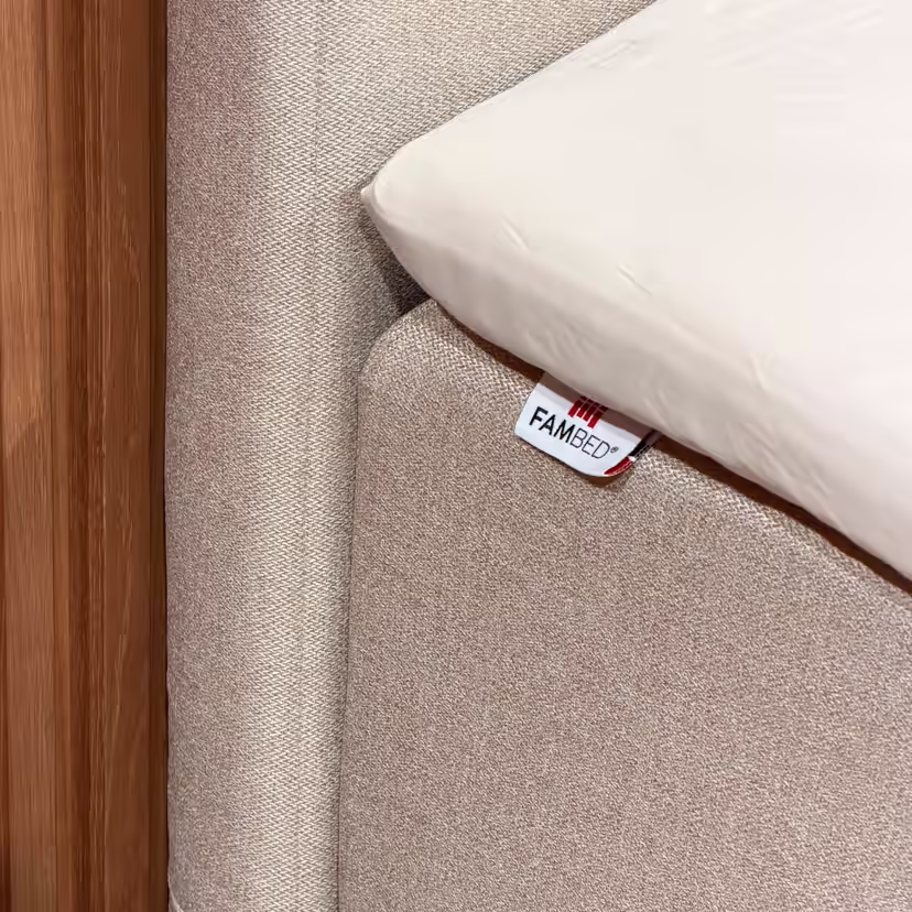 Nærbilde av det beige trekket rundt sengens hjørne, med FAMBED-merke og hvitt laken oppå